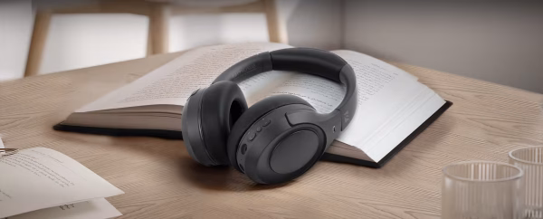 Le cuffie M-295 ANC sono mostrate ripiegate e inserite a metà nella loro custodia da viaggio nera con zip, accanto a un passaporto e un libro, per sottolinearne la vocazione da viaggio."
